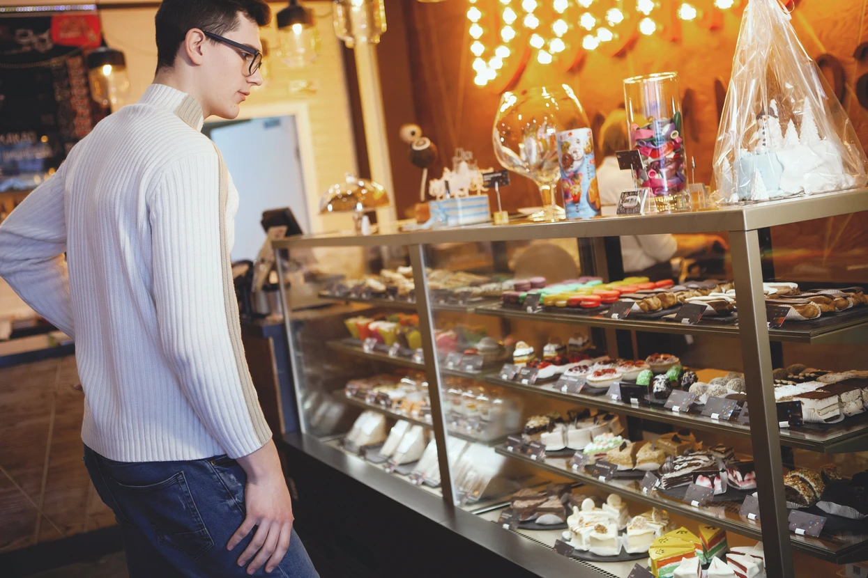 Jeune homme en pull beige et lunettes examine une vitrine réfrigérée de chocolaterie artisanale