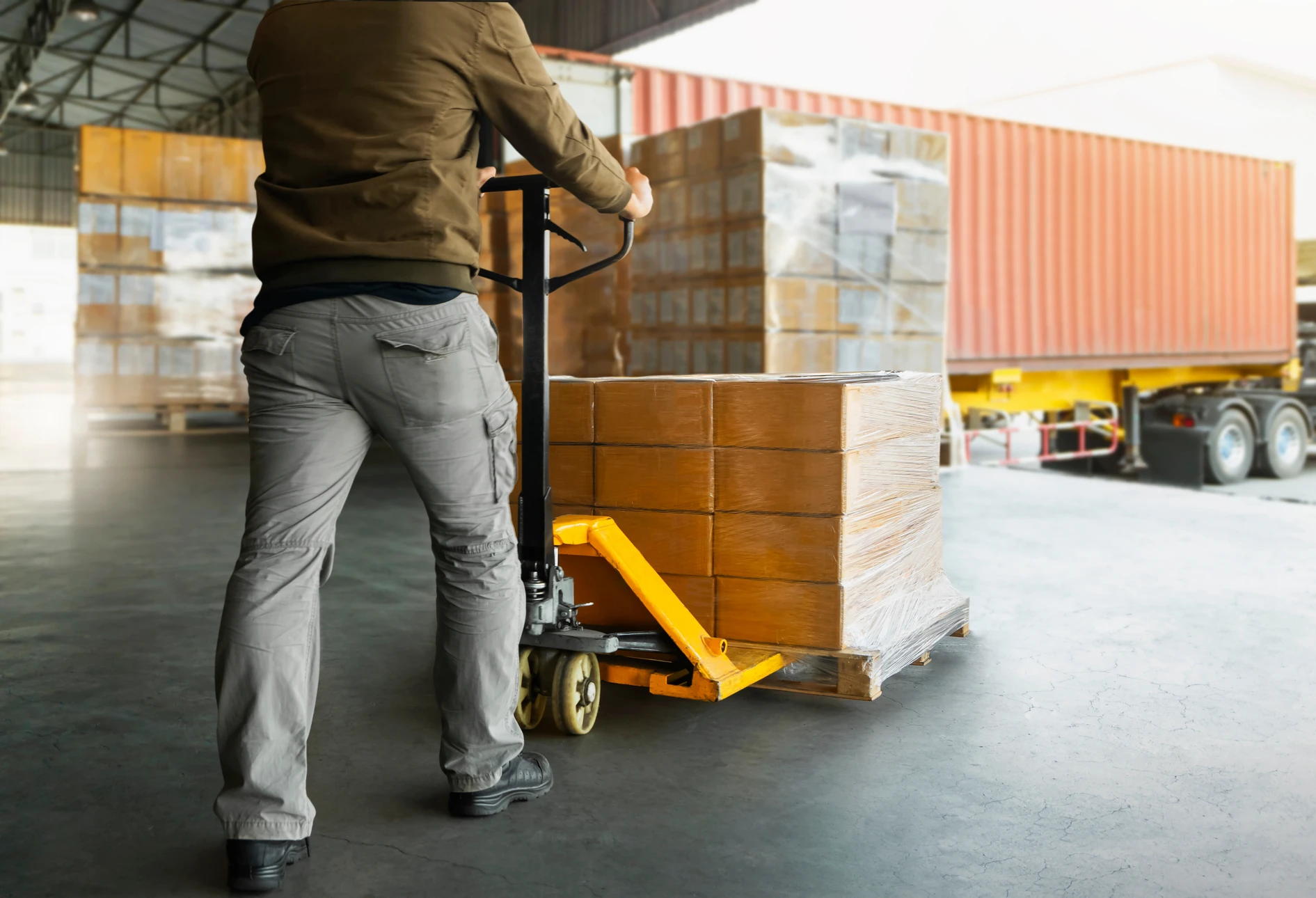 Un employé d'entrepôt manœuvre un transpalette manuel dans un centre logistique, transportant des colis emballés vers un camion de livraison