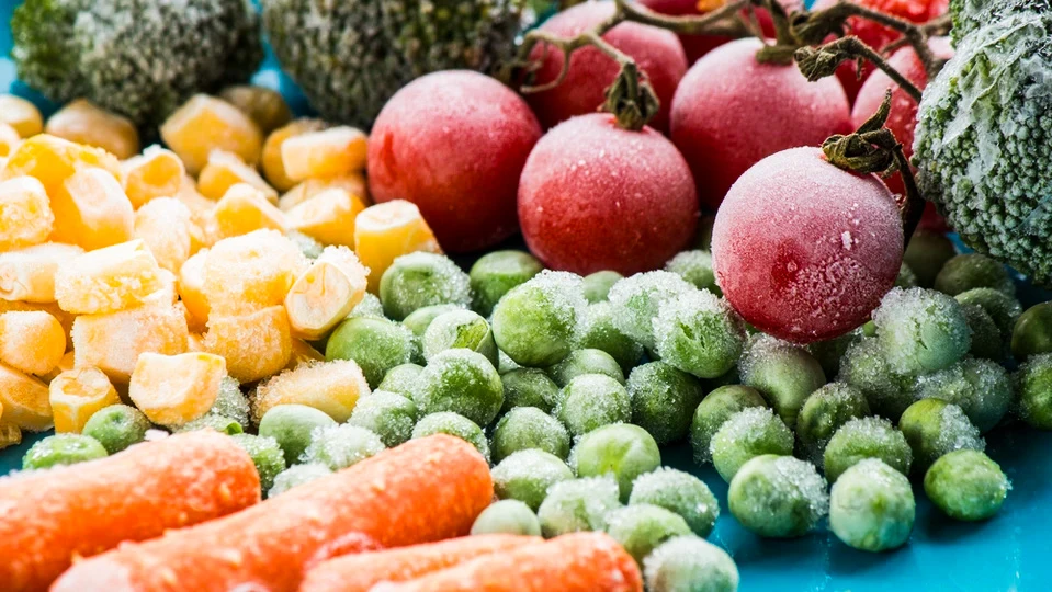 Assortiment de légumes surgelés disposés sur surface bleue : petits pois verts givrés, maïs en grains dorés, radis cherry rouges avec tiges, brocolis verts et carottes orange coupées en bâtonnets